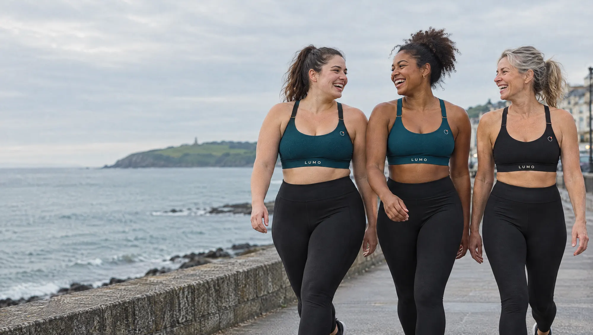 Three women of different ages and body sizes laughing as they walk along a Dublin seafront, wearing The Lift in Deep Sea and Ink.